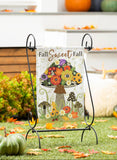 Decorative garden flag with floral design and 'Fall Sweet Fall' text on a stand, surrounded by pumpkins and autumn leaves.