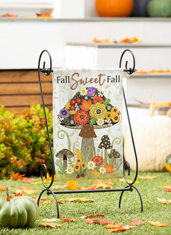 Decorative garden flag with floral design and 'Fall Sweet Fall' text on a stand, surrounded by pumpkins and autumn leaves.
