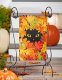 Decorative flag with a black cat and autumn leaves on an easel, surrounded by pumpkins and fall foliage.