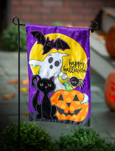 Halloween-themed garden flag with a ghost, black cat, and pumpkin in front of pumpkins.