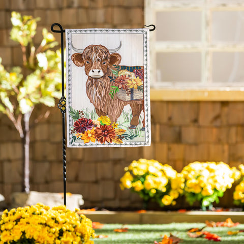 Decorative garden flag with a cow and flowers on a stand in front of yellow flowers.