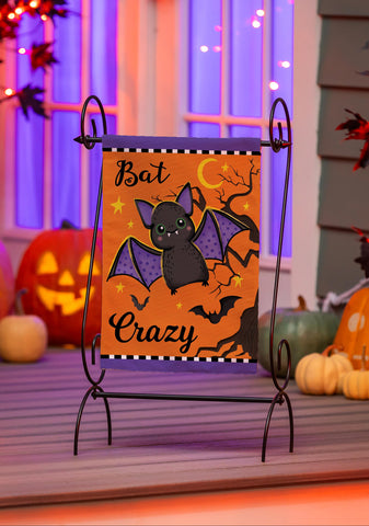 Halloween-themed decorative sign with a bat illustration on an easel, surrounded by pumpkins and festive lights.