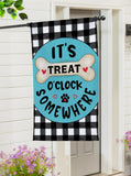 Decorative flag with 'It's Treat O'Clock Somewhere' text on a black and white checkered background, hanging outside a house.