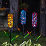 Three colorful glass flower lights on stakes with a blurred garden background.
