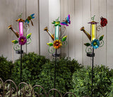 Decorative solar lights shaped like watering cans with colorful flowers and butterflies in a garden setting.