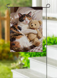 Cat with a teddy bear on a garden flag held by a stand outdoors.