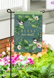 Decorative garden flag with birds and floral design on a stand in front of flowers and plants.