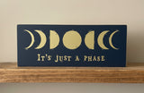 Decorative sign with moon phases and text 'It's Just a Phase' on a wooden shelf.