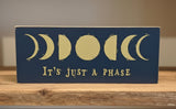 Decorative sign with moon phases and text 'It's Just a Phase' on a blue background.